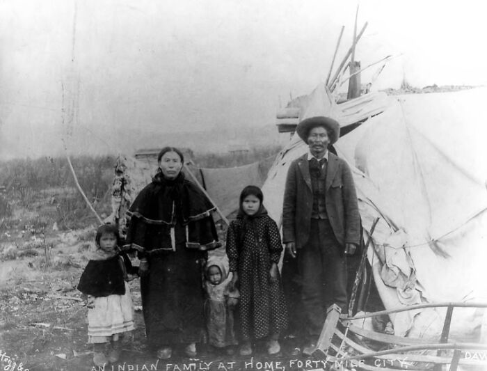 Vintage Native American family standing outside a traditional tipi, dressed in early 20th-century clothing.