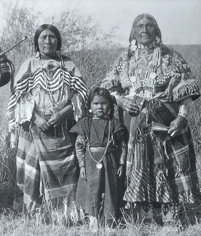 Vintage Native American family dressed in traditional clothing standing outdoors in a natural grassy setting.