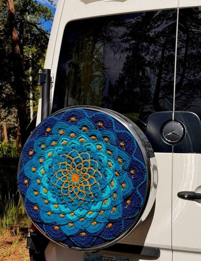 Crocheted blue and orange mandala pattern covering a spare tire on a white Mercedes vehicle outdoors.
