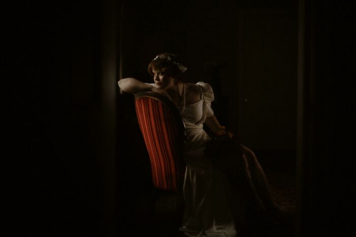 Woman in a white dress posing on a red chair in low light creating an accidental Renaissance photo effect.
