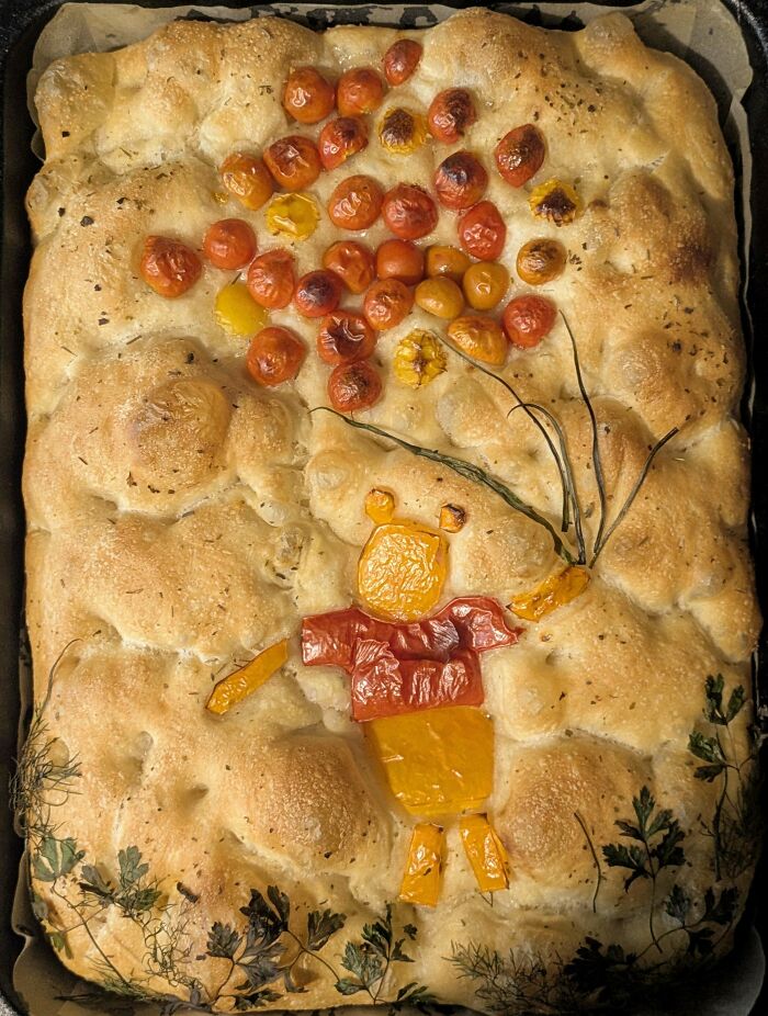 Baked focaccia bread decorated with vegetables forming a playful figure holding balloon-shaped tomatoes, showcasing gorgeous baked goods.