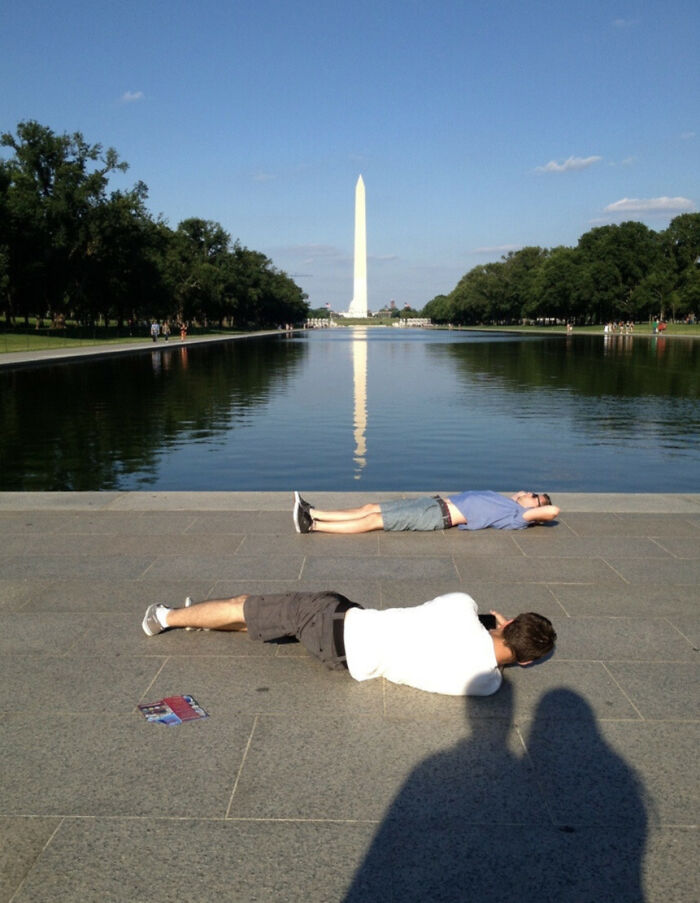 Man taking a creative vacation pic lying on ground with Washington Monument reflected in water behind him.