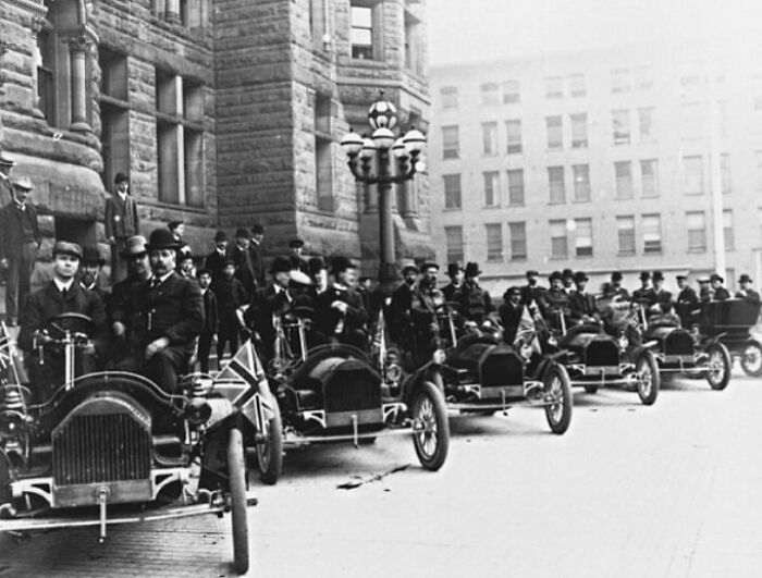 Row of wild first cars from 100 years ago with drivers in early 20th-century attire in a city street setting.