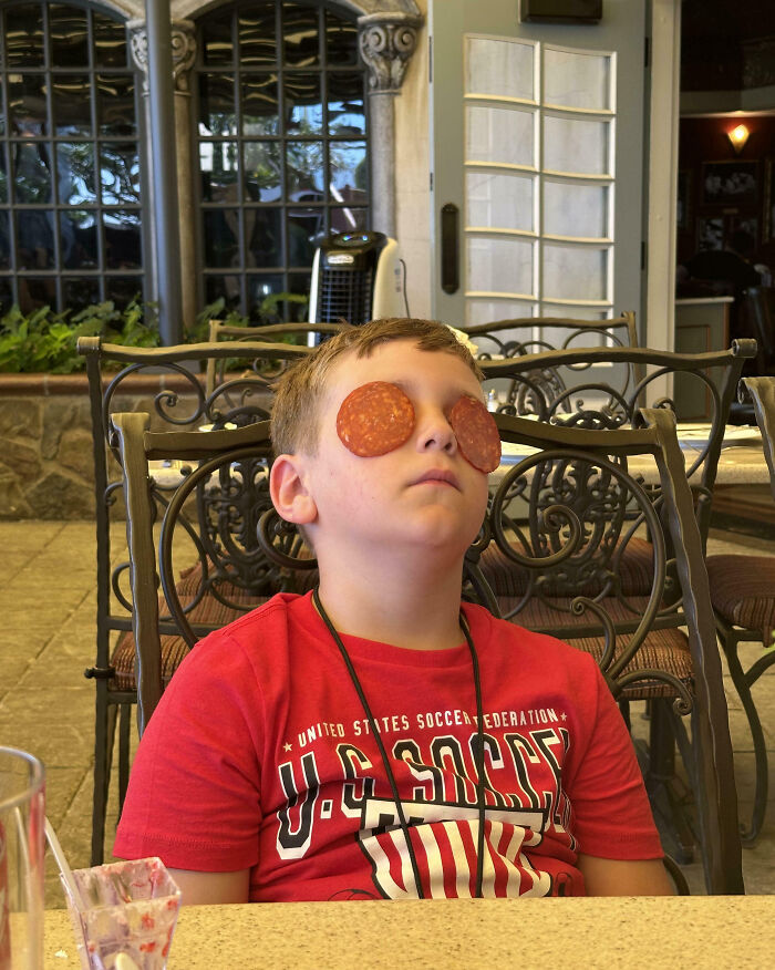 Boy on vacation with pepperoni slices over his eyes, sitting outdoors at a table in a humorous vacation pic moment.