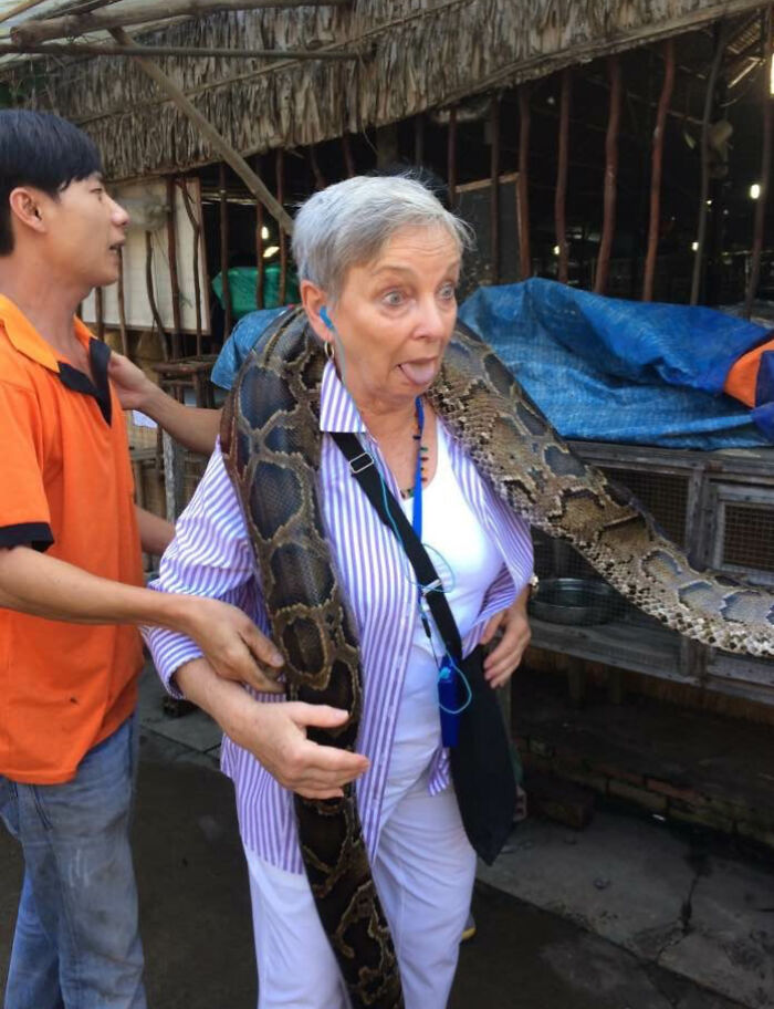 Older woman making a funny face while holding a large snake on vacation, capturing a moment fit for comedy special.