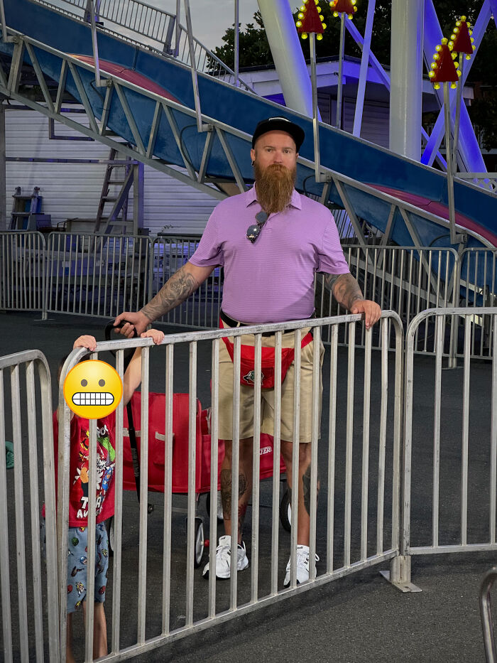 Man with a long beard and tattoos stands behind metal barriers at carnival, funny vacation pics with child hiding behind bars.