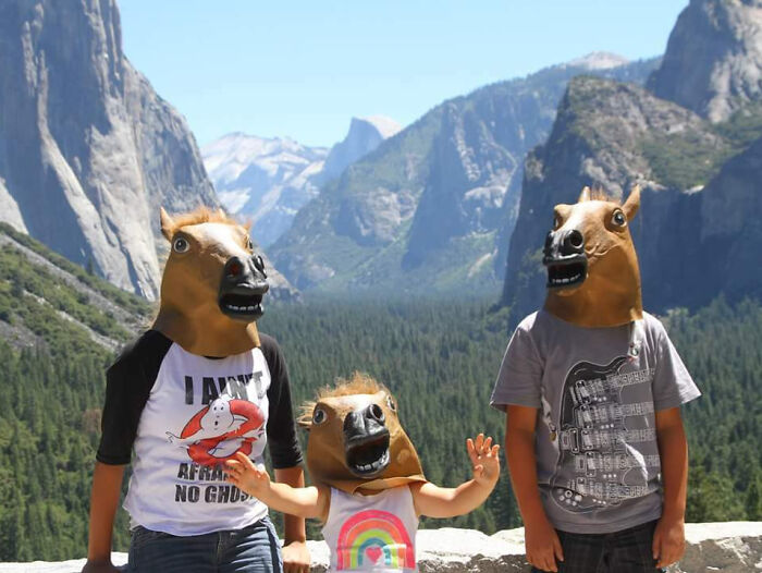 Three people wearing horse masks pose for a funny vacation pic with mountains and forest in the background.