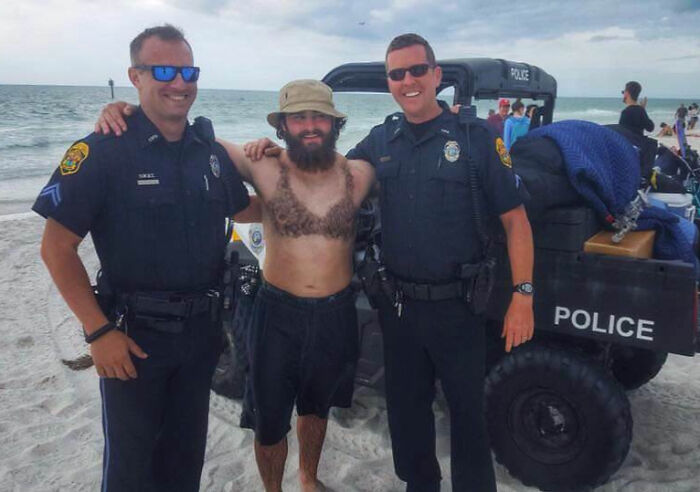 Man with fake chest hair posing with two smiling police officers on a beach, a funny vacation pic moment captured.