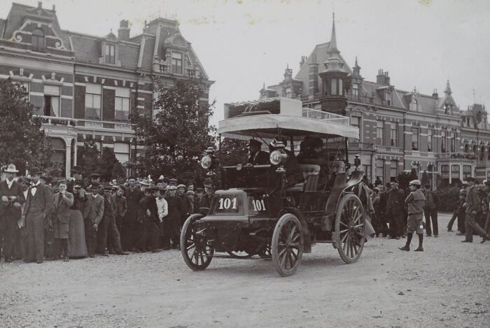 Vintage photo of a wild first car from 100 years ago surrounded by a crowd in an early 20th-century city setting