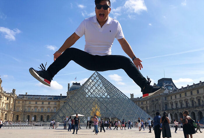 Man wearing sunglasses jumping with legs spread over Louvre Pyramid, a funny vacation pic at a famous Paris landmark.