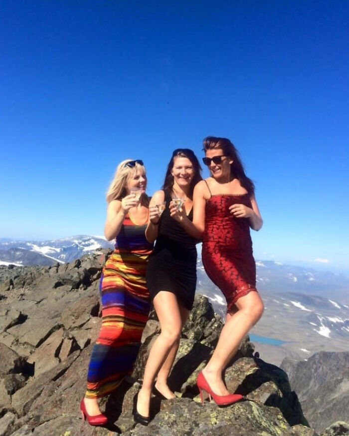 Three women wearing dresses and high heels posing on rocky mountain top in a hilarious vacation pics moment.