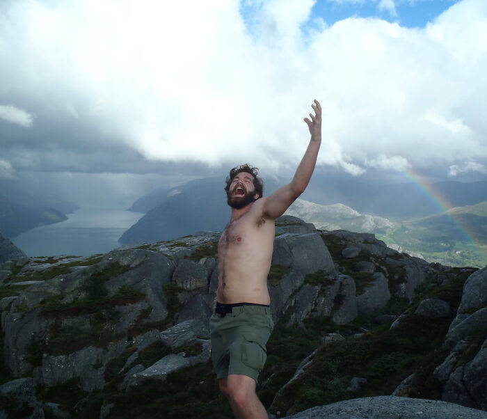 Shirtless man posing dramatically on rocky mountain during vacation with a rainbow in the cloudy sky, a funny vacation pic.
