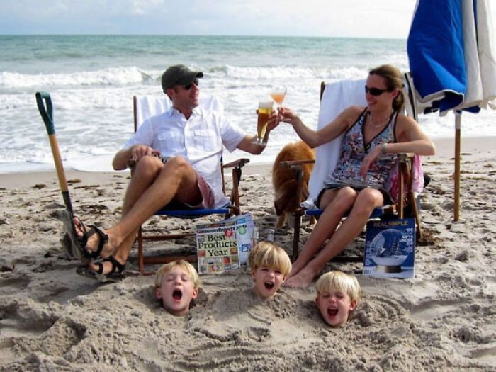 Couple toasting drinks on beach chairs while three kids buried in sand scream, a funny vacation pics moment.