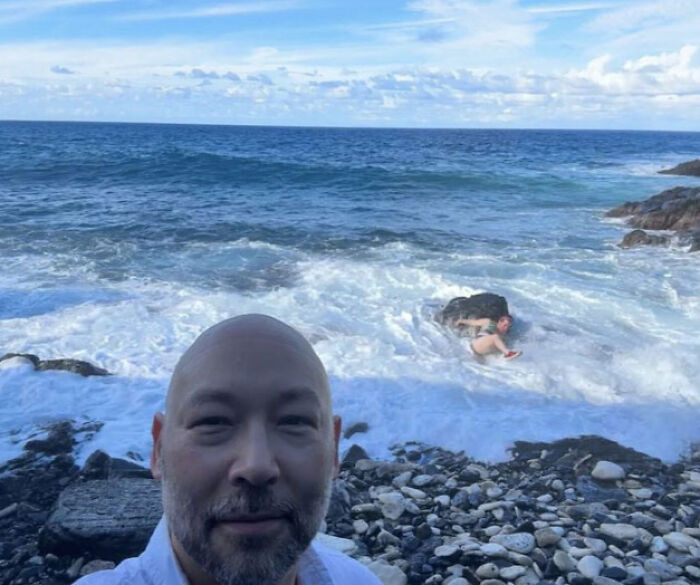 Man takes vacation pic on rocky shore as another person struggles in ocean waves in a funny vacation pics moment.