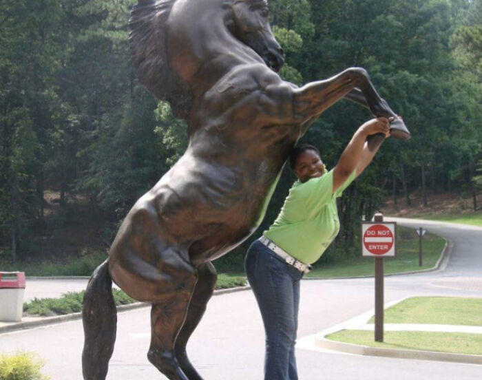 Person posing humorously with a rearing horse statue, creating a vacation pic perfect for a comedy special moment.