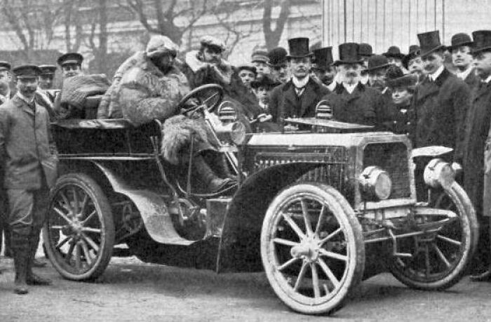 Early 20th-century first cars showcased with people in period clothing around the vintage vehicle outdoors.