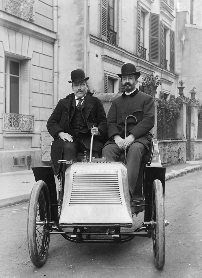 Two men in bowler hats sitting in a wild first car from 100 years ago on a street in a vintage black and white photo