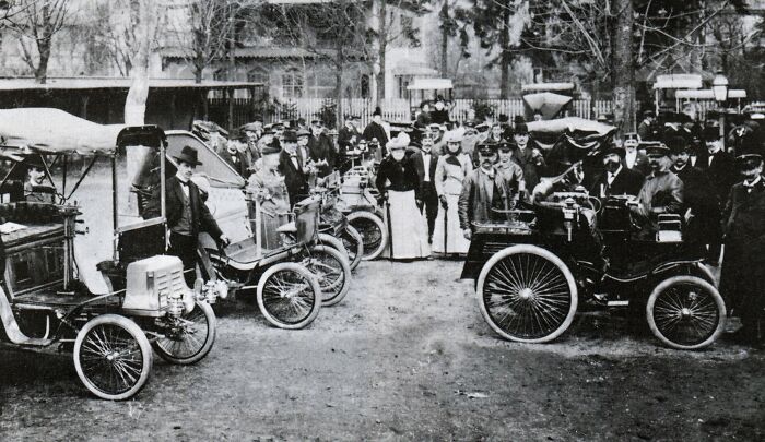 Crowd of people gathered around wild first cars from 100 years ago showcasing early automotive design and innovation.