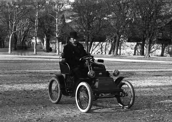 Man in early 1900s clothing driving a wild first car on a dirt road surrounded by leafless trees.