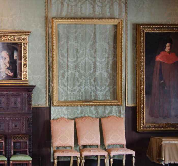 Empty antique gold frame hanging on a vintage wall with classic paintings and three upholstered chairs below, mystery theme