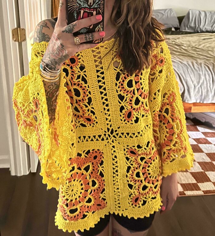 Woman showing a yellow and orange crocheted top with floral patterns in a casual indoor setting.