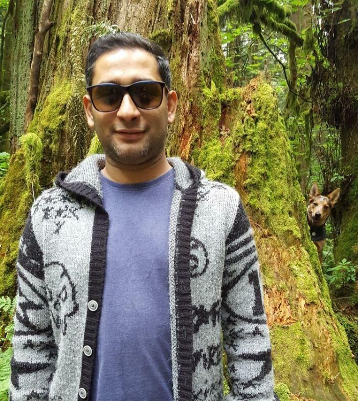 Man wearing sunglasses posing in forest while dog funny photo bombs behind moss-covered tree, showcasing pets crashing photos.