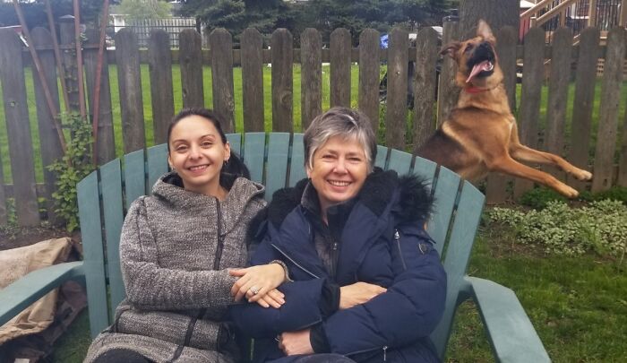 Two women smiling on a bench while a dog photobombs by leaping in the air, showcasing funny times pets crashed photos.