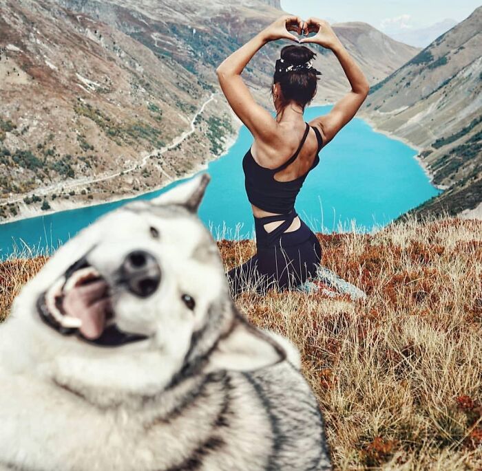 Smiling dog photobombs woman posing by blue mountain lake, showcasing funny times pets crashed photos and stole the show.