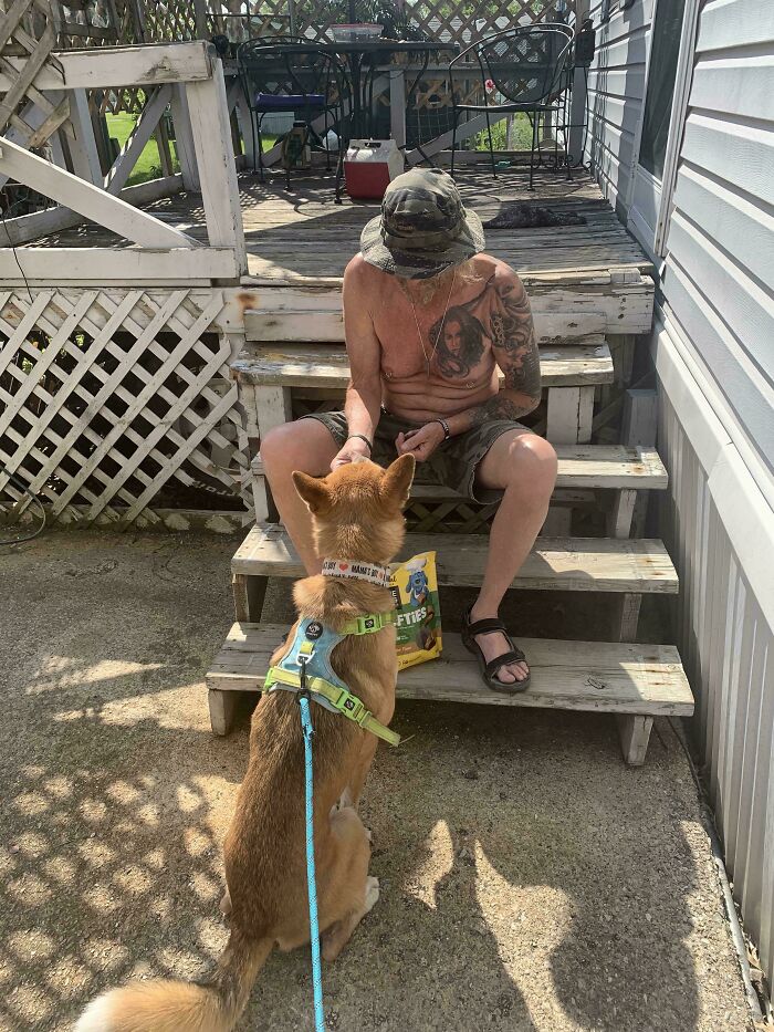 Man with tattoos sitting on wooden steps giving treats to a dog on leash outside, showcasing wholesome kind neighbors bond.