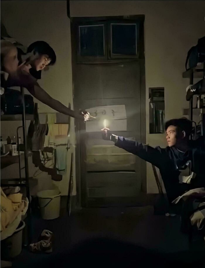 Two men in a dim room exchanging light from a candle and match, resembling an accidental Renaissance photo moment.