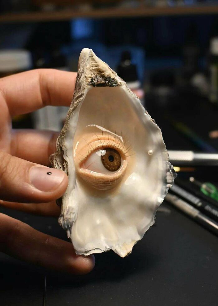 Hand holding an oyster shell with a realistic human eye inside, showcasing examples of awful taste but great e*******n.