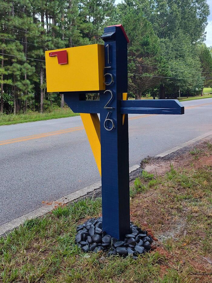 Woodworker’s creative mailbox project painted yellow and navy blue with house numbers 1526, standing by roadside.