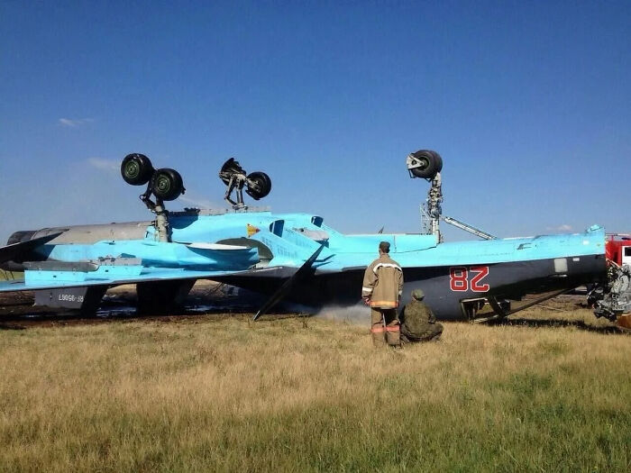 Upside-down military aircraft on grass with two people inspecting it, showcasing a dramatic parking fail scene.