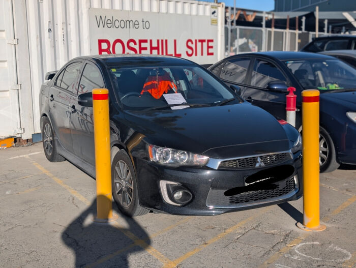 Black car parked incorrectly between yellow bollards with a parking ticket on the windshield at a construction site.