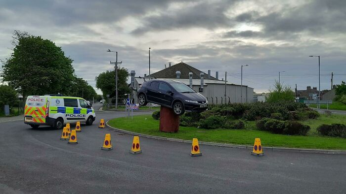 Car stuck on a roundabout pillar with police van and traffic cones nearby illustrating a parking fail.