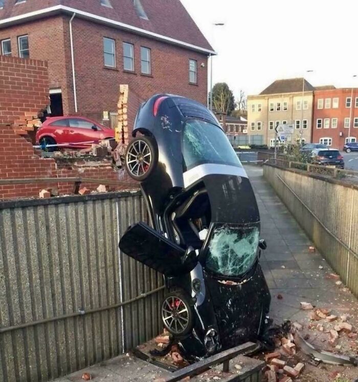 Black car crashed vertically against fence after parking fail, broken windows and damaged brick wall visible in urban area.