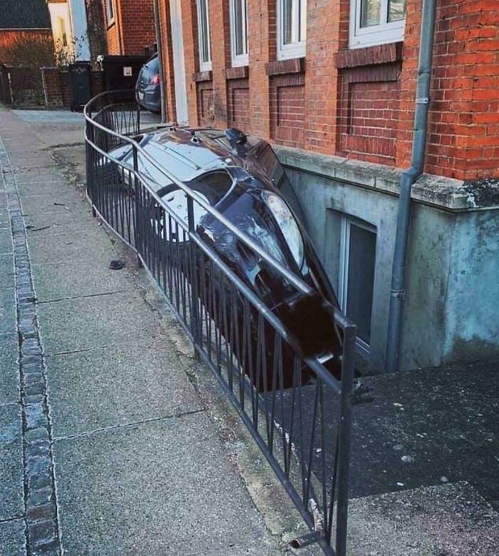 Black car crashed and stuck between metal railing and building wall in an unusual parking fail on a sidewalk area.