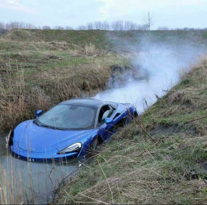 Blue sports car stuck in a narrow water ditch, surrounded by grass and smoke rising nearby, showcasing a major parking fail.
