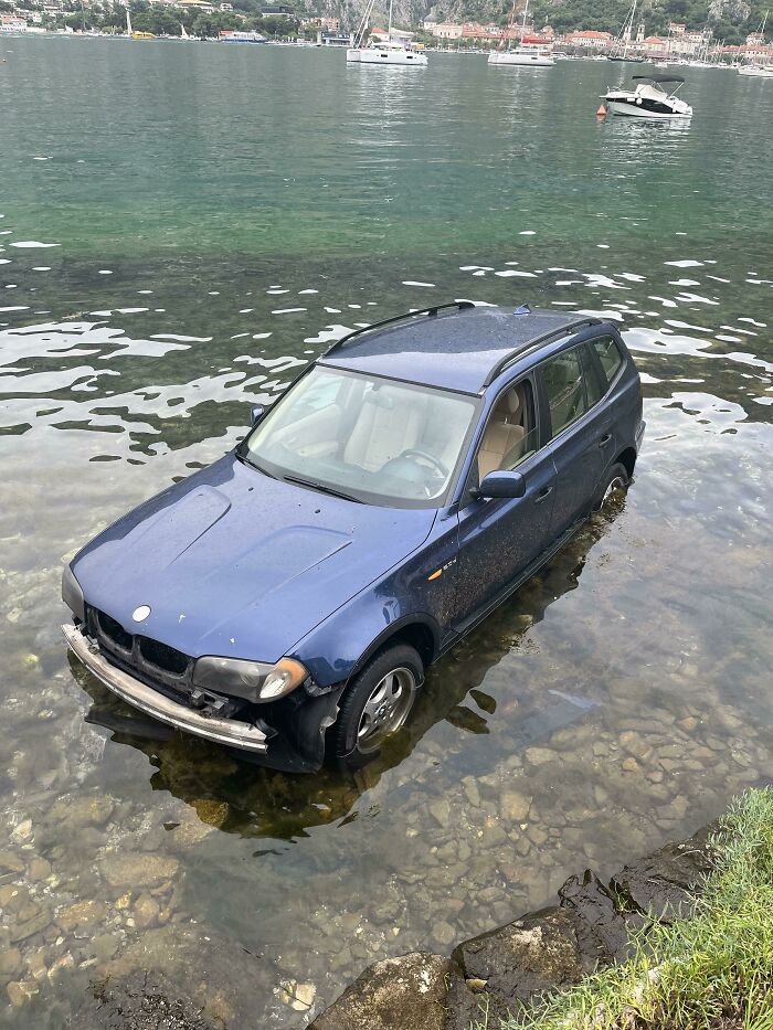 Blue SUV stuck in shallow water near rocky shore, illustrating a major parking fail shared online.