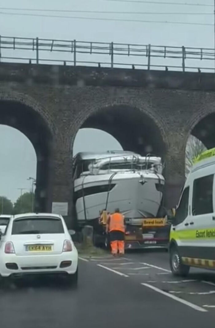 Large boat stuck under low bridge on road, causing a notable parking fail with vehicles nearby and a worker in high-visibility clothing.