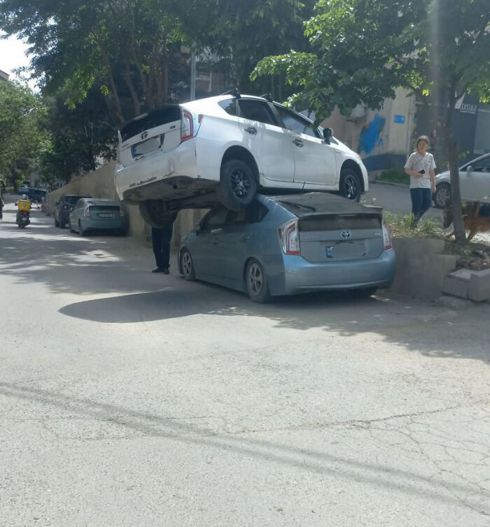 Two Toyota Prius cars involved in a parking fail, one parked on top of the other on a city street.