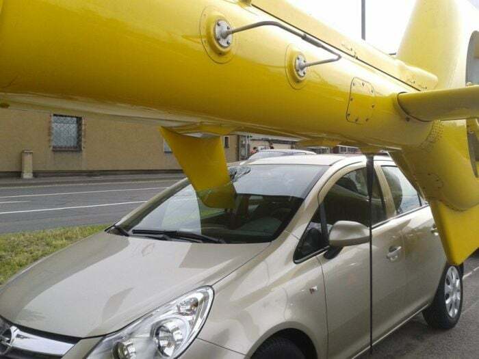 Small car awkwardly parked under a yellow airplane wing, illustrating a funny parking fail shared online.