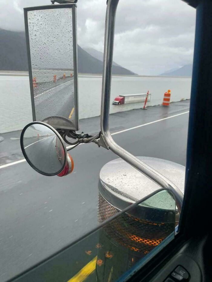 View from truck mirror showing a semi-truck trailer fallen off the road near a body of water, a clear parking fail scene.