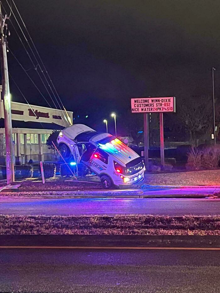 Police car stuck partially upright next to a utility pole at night, showcasing a serious parking fail with flashing lights.