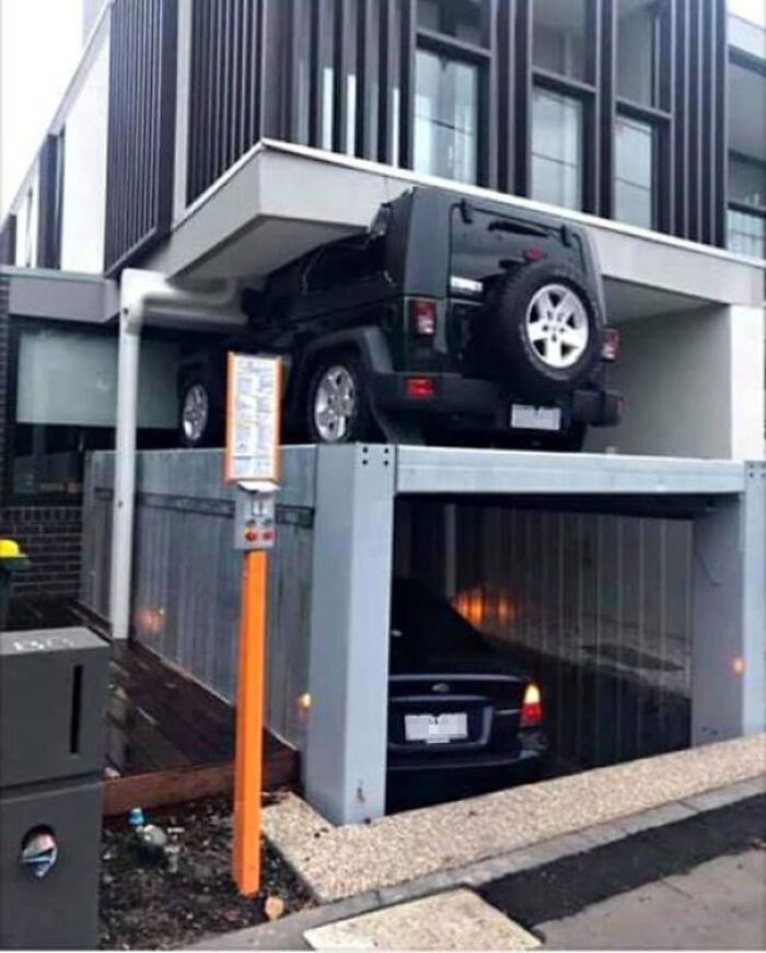 Black Jeep parked awkwardly on a narrow overhead platform above a small car in a tight parking fail spot.