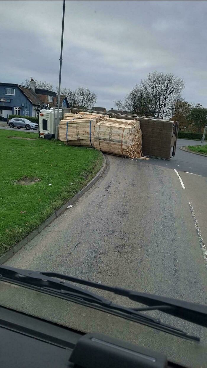 Truck tipped over on curved road, spilling wooden planks and causing a major parking fail on a cloudy day.