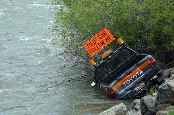 Pickup truck stuck in water by riverbank with a pilot car follow me sign, showcasing a parking fail.