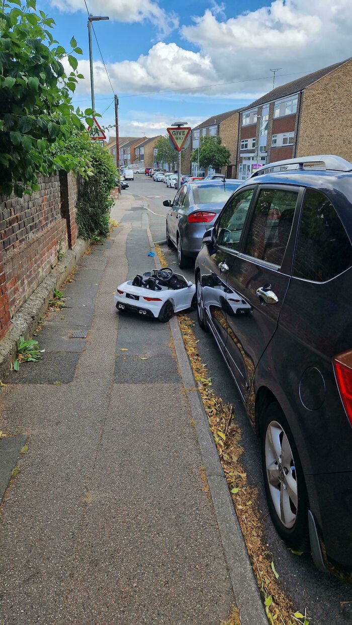 Small white toy car parked on sidewalk next to full-sized cars, illustrating a funny parking fail on a residential street.