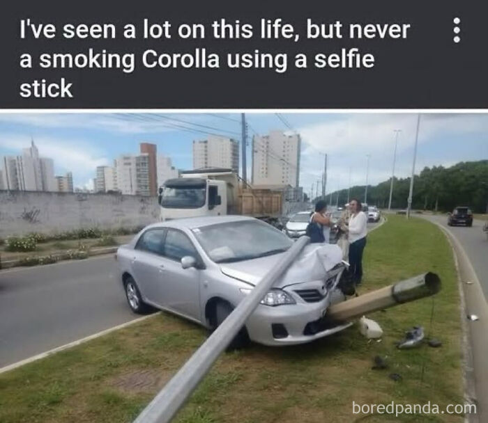 Silver car crashes into a streetlight on grass median, illustrating a parking fail on a busy urban road with bystanders nearby.