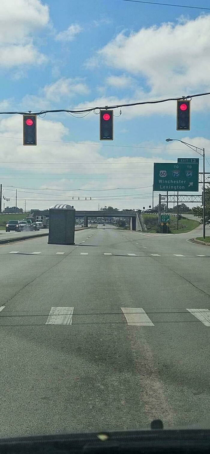 Portable toilet abandoned in the middle of the road causing a parking fail on a clear day with red light signals ahead.